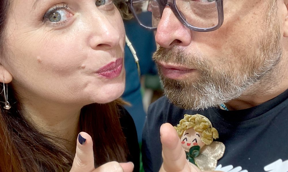 Due persone sorridenti indicano verso la fotocamera